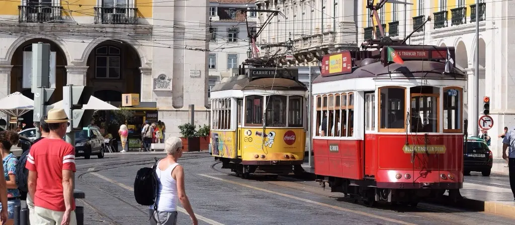 Yellow Carris tram and red Lisbon Tram Tour at Praça do Comércio, with tourists crossing the cobblestone square