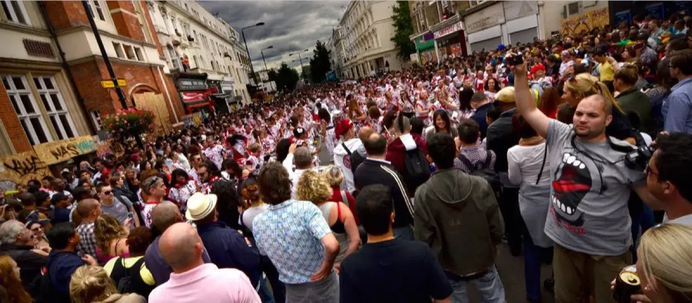 Dense crowd of spectators and white-and-red costumed performers filling a terraced London street at Notting Hill Carnival under an overcast sky