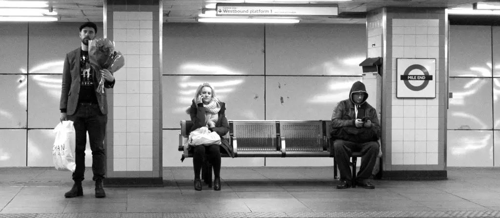 Black-and-white shot of three figures on Mile End Underground platform: a man with flowers, a seated woman, a hooded figure