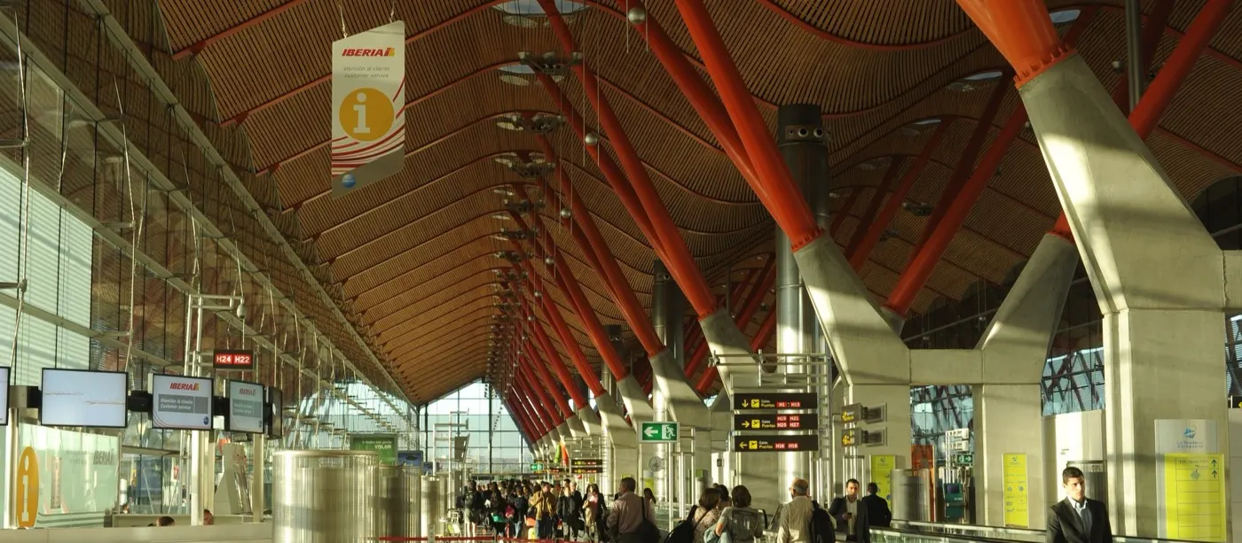Madrid-Barajas Terminal 4 hall with red steel columns under a wooden ceiling, an Iberia customer service desk on the left, and passengers walking towards the gates