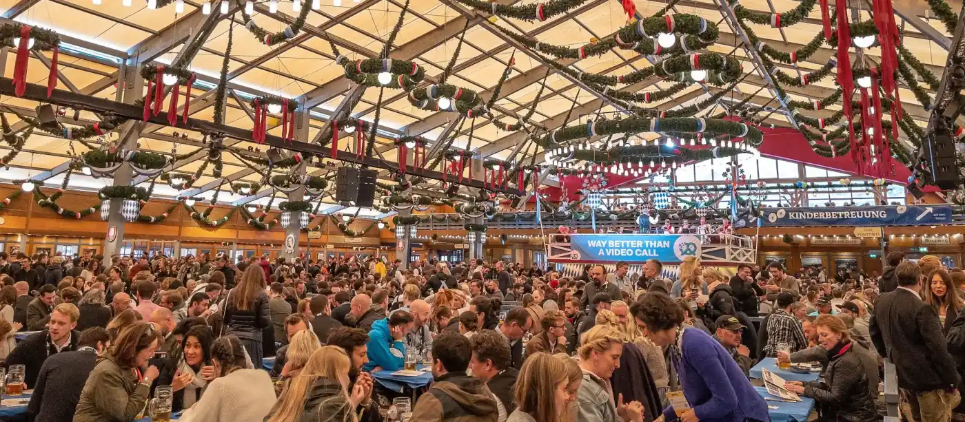 Packed Oktoberfest beer tent interior with pine garlands and red ribbons hanging from the roof trusses and a Kinderbetreuung childcare sign above the crowd