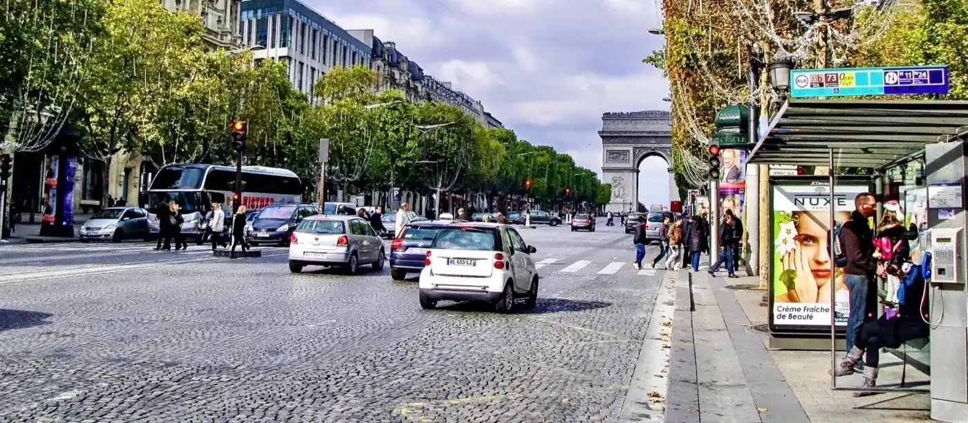 Champs-Élysées avenue in Paris with a bus stop shelter and route signage in the foreground, cars and a tour coach on the road, and the Arc de Triomphe visible in the distance