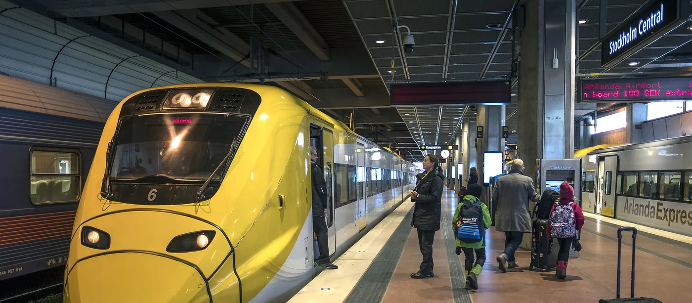 Yellow Arlanda Express train stopped at a Stockholm Central platform with passengers and rolling luggage walking toward it and another Arlanda Express carriage on the opposite track
