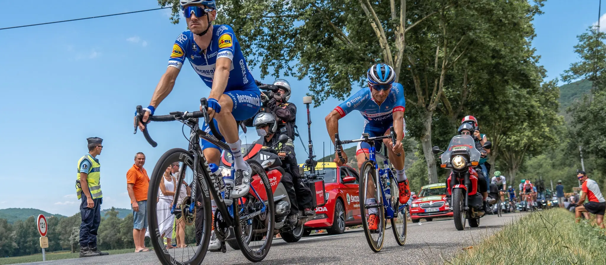 Two Tour de France riders climbing a tree-lined road past spectators, with police, motorbikes and support cars trailing behind