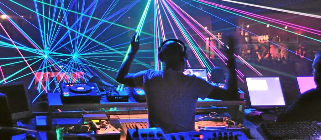 Silhouetted DJ at the decks with arms raised, surrounded by sweeping green and pink laser beams